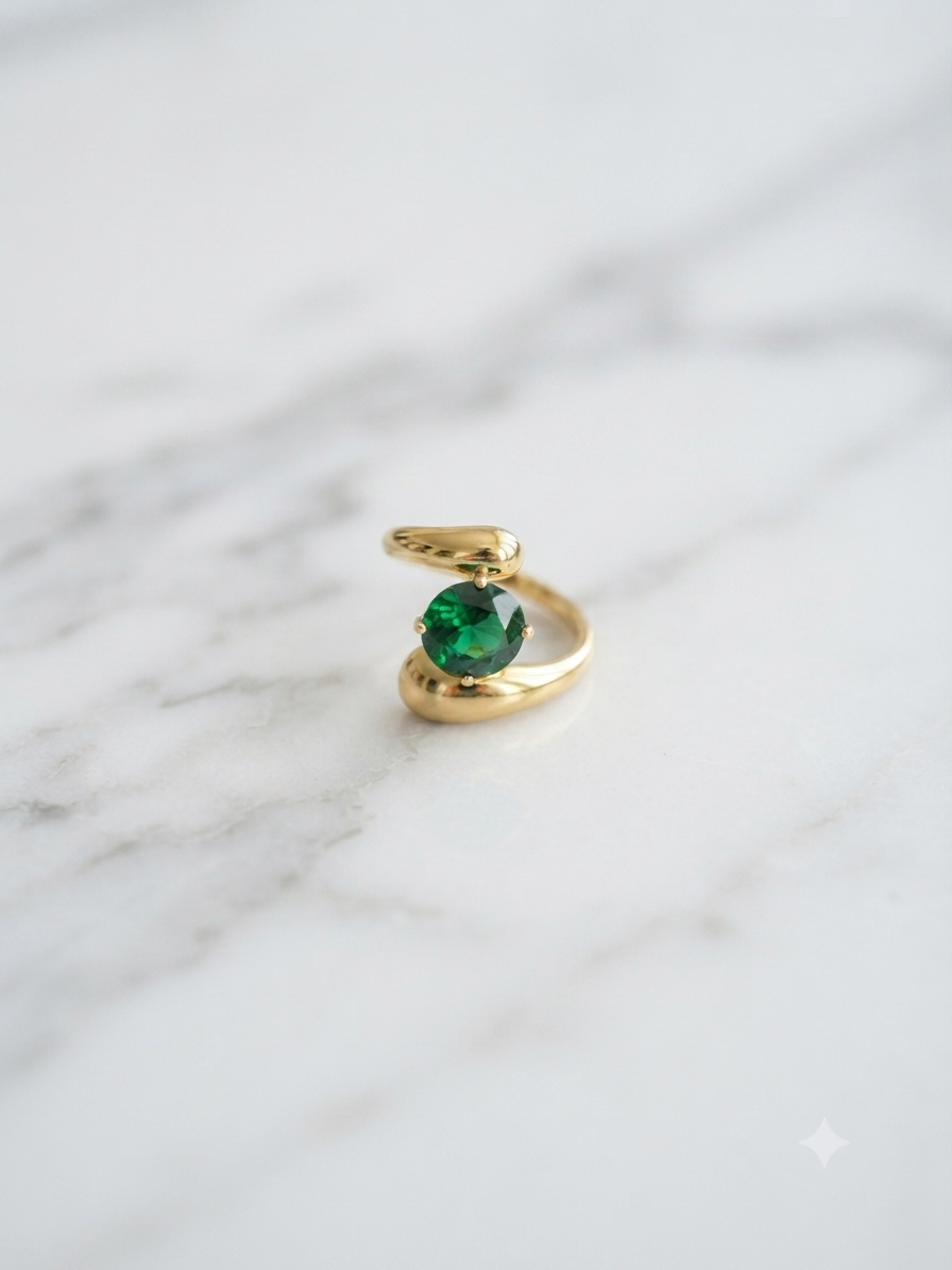 Bague ajustable dorée en acier inoxydable avec pierre vert émeraude, posée sur fond marbre blanc.