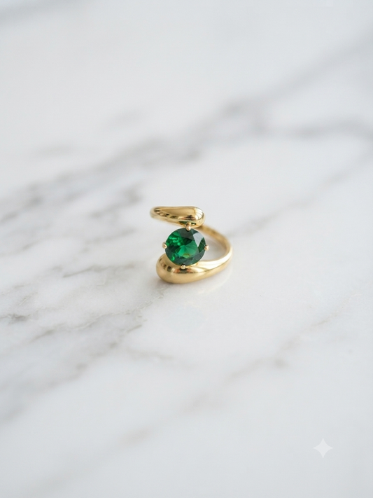 Bague ajustable dorée en acier inoxydable avec pierre vert émeraude, posée sur fond marbre blanc.